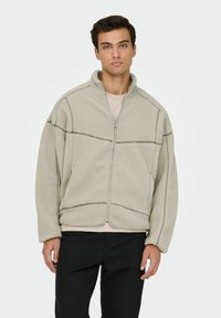 Giacca in pile color beige chiaro con colletto alzato, cuciture a contrasto, chiusura frontale con zip e tasche laterali. Indossata sopra una camicia color crema.