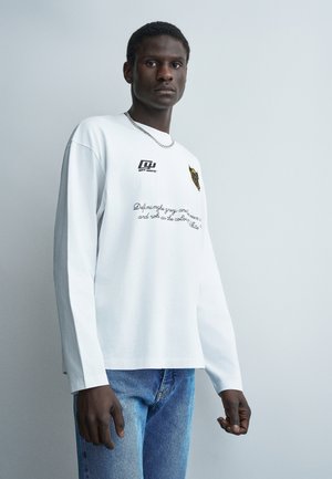 Muž v bielom tričku s dlhým rukávom s textom a logom, modrých džínsoch, striebornej retiazke na krku, stojaci pred jednoduchým svetlošedým pozadím.