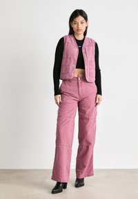 Gilet en coton rose et pantalon large assorti, avec un devant à boutons, des poches rabat et un tissu texturé. Haut ajusté noir et bottes.