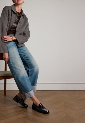 Zwarte leren instappers met een gladde textuur, gedragen met lichtblauwe denim jeans met opgerolde pijpen en een grijze oversized jas, zittend op een houten stoel.