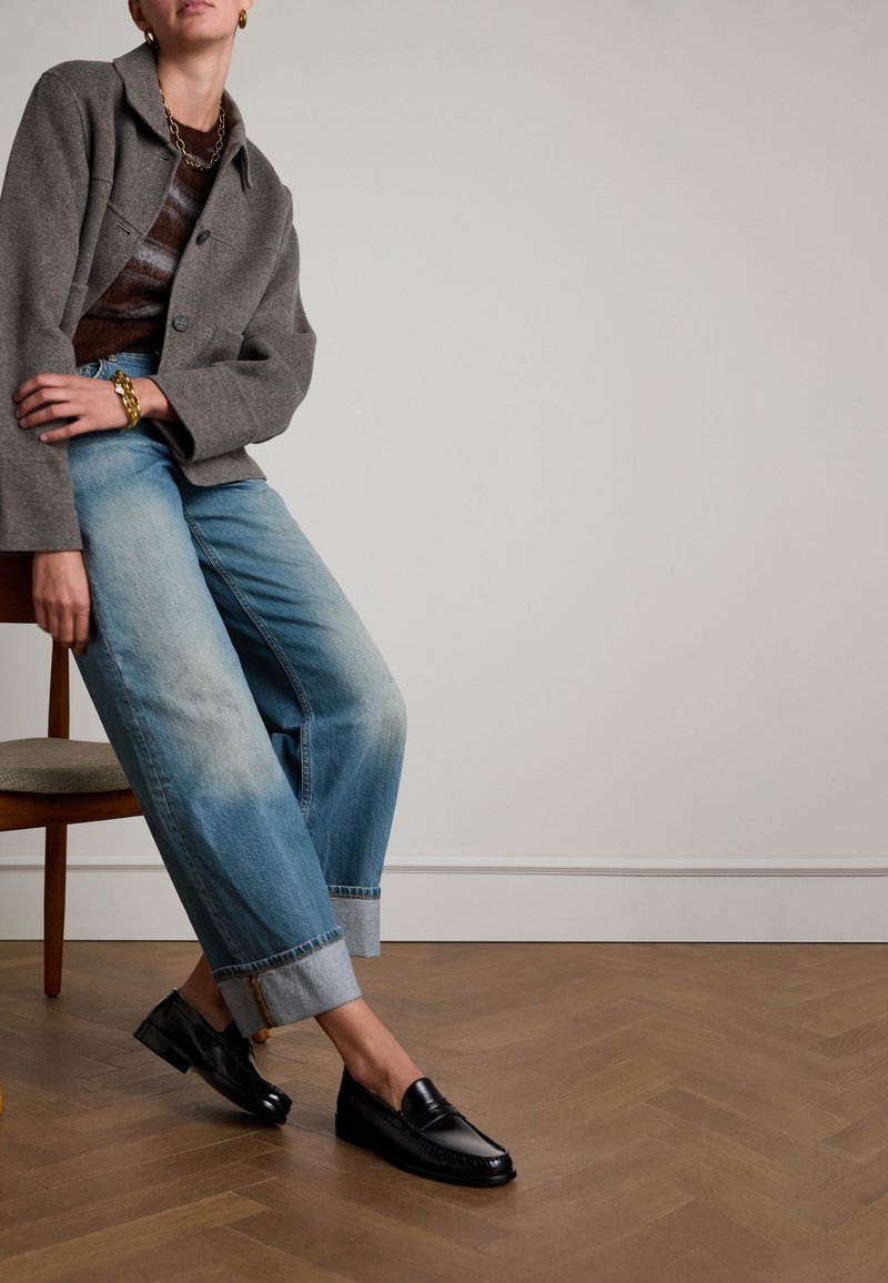 Schwarze Lederloafers mit glatter Textur, getragen mit hochgekrempelten hellblauen Denim-Jeans und einer grauen übergroßen Jacke, sitzend auf einem Holzstuhl.