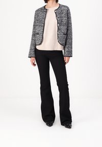 Giacca nera e bianca con texture, collo rotondo e bottoni argentati, indossata sopra un maglione beige chiaro e abbinata a pantaloni neri a zampa d'elefante.