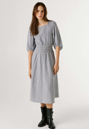 Femme portant une robe à rayures verticales grises et blanches avec des manches bouffantes et des bottes noires mi-mollet à boucle, debout devant un fond uni.