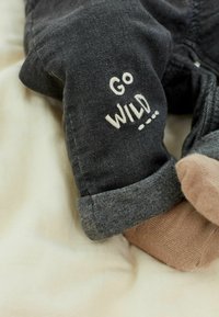 Pantalon gris foncé avec le texte brodé "Go Wild" sur la jambe, porté avec des chaussettes marron sur une surface en tissu clair.