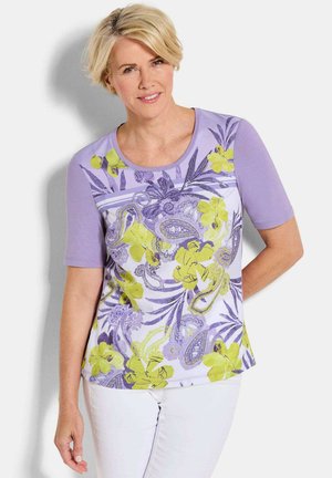 Femme aux cheveux blonds courts, portant un haut violet à manches courtes à motif floral et un pantalon blanc, debout sur un fond clair.