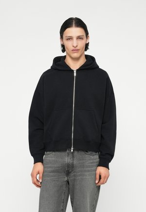 Sweatshirt à capuche noir avec fermeture éclair à l'avant, présentant un design court, de grandes poches avant et des poignets côtelés. Tissu doux au toucher lisse.