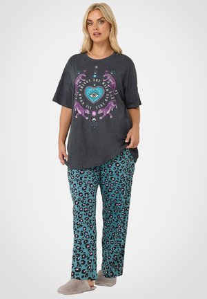 Femme portant un T-shirt large gris foncé avec des léopards violets et un motif cœur bleu, un pantalon imprimé léopard bleu-vert et des chaussons gris, debout.