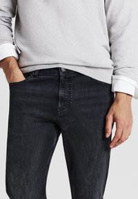 Maglione grigio con polsini bianchi, abbinato a jeans di denim scuri. I jeans hanno una vestibilità classica e una texture sottile. Le mani sono in tasca.