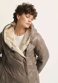 Kurtka pikowana w kolorze taupe z kremowym podszewką, z dużym kapturem, ukośnym przeszyciem i zapięciem na zatrzaski przy dekolcie.