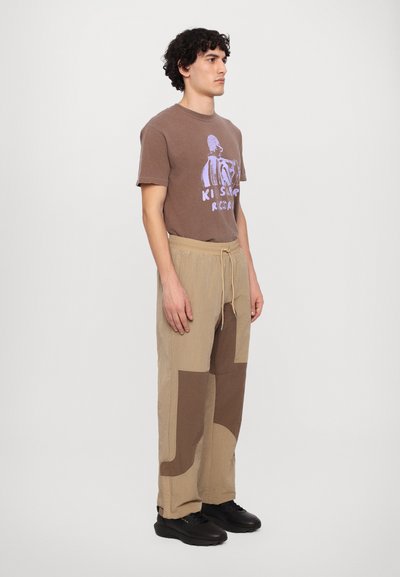 Brun grafisk t-shirt med et lilla design, parret med beige drawstring-bukser med store brune lapper, og sorte sneakers.