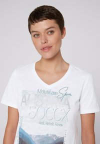 Biała koszulka z dekoltem w serek z graficznym nadrukiem zawierającym tekst "Austria" oraz "Mountain Spa", podkreślona malowniczym tłem gór.