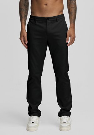 Uomo che indossa pantaloni neri aderenti e scarpe da ginnastica bianche, in piedi davanti a uno sfondo grigio uniforme con tatuaggi visibili sugli avambracci.