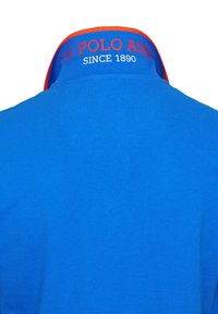 Modra polo majica z rdečim ovratnikom, na katerem je s bele niti vezeno "U.S. POLO ASSN" in "SINCE 1890". Gladka tekstura tkanine, klasičen dizajn.