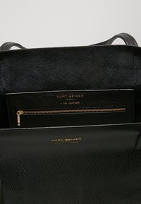 Svart läder tote-väska med en guldfärgad dragkedjeficka inuti. Har en mjuk, texturerad insida, subtil märkning och dubbla handtag.