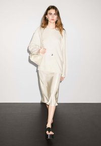 Maglione crema con maniche voluminose, gonna beige in stile satin, sandali neri con plateau e una piccola borsa chiara. Linee semplici e pulite.