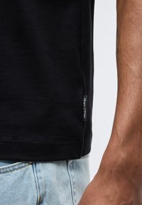 Μαύρη άκρη πουκάμισου με μικρή ετικέτα «Marc O'Polo» φορεμένη πάνω από ανοιχτόχρωμο τζιν, μερική θέα μπράτσου με τατουάζ.