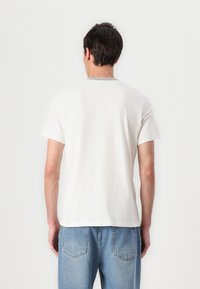 Tricou alb cu mânecă scurtă, cu guler rotund și cusături de accent verde. Țesătura pare moale, cu o croială lejeră și detalii minime.
