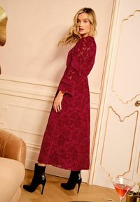 Burgundy kukkais pitsimekko, jossa on röyhelöedestä ja tiukka vyötärö, yhdistetty mustiin stiletto nilkkureihin. Rakenteellinen ja elegantti muotoilu.