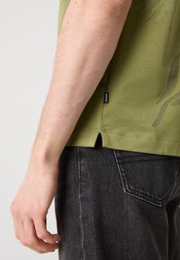 Kratke rokave, olivno zelena srajca iz teksturirane tkanine, z stranskimi razpoki na robu in črno oznako. V kombinaciji s temno denim jeansami.