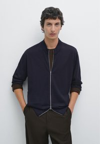 Námořnický svetr na zip s vysokým límcem, vyrobený z měkkého materiálu. Má rozdělený lem a je spojen s hnědými kalhotami. Jednoduchý design, žádné viditelné vzory.