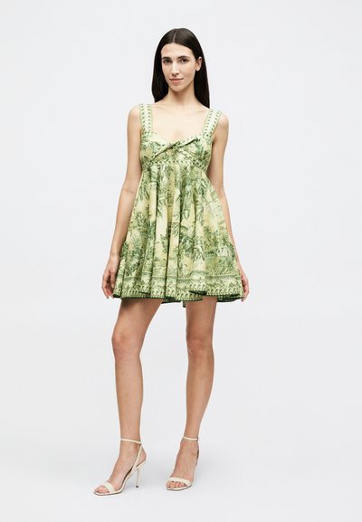 Γυναίκα με μακριά σκούρα μαλλιά, ντυμένη με πράσινο μίνι φόρεμα χωρίς μανίκια με τροπικό σχέδιο και μπεζ ψηλοτάκουνα σανδάλια με λουράκια, στέκεται μπροστά σε λευκό φόντο.