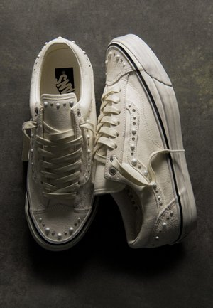 Чифт мръснобели платнени кецове Vans с връзки, метални капси и гумени подметки с черни ивици, подредени върху тъмна повърхност.