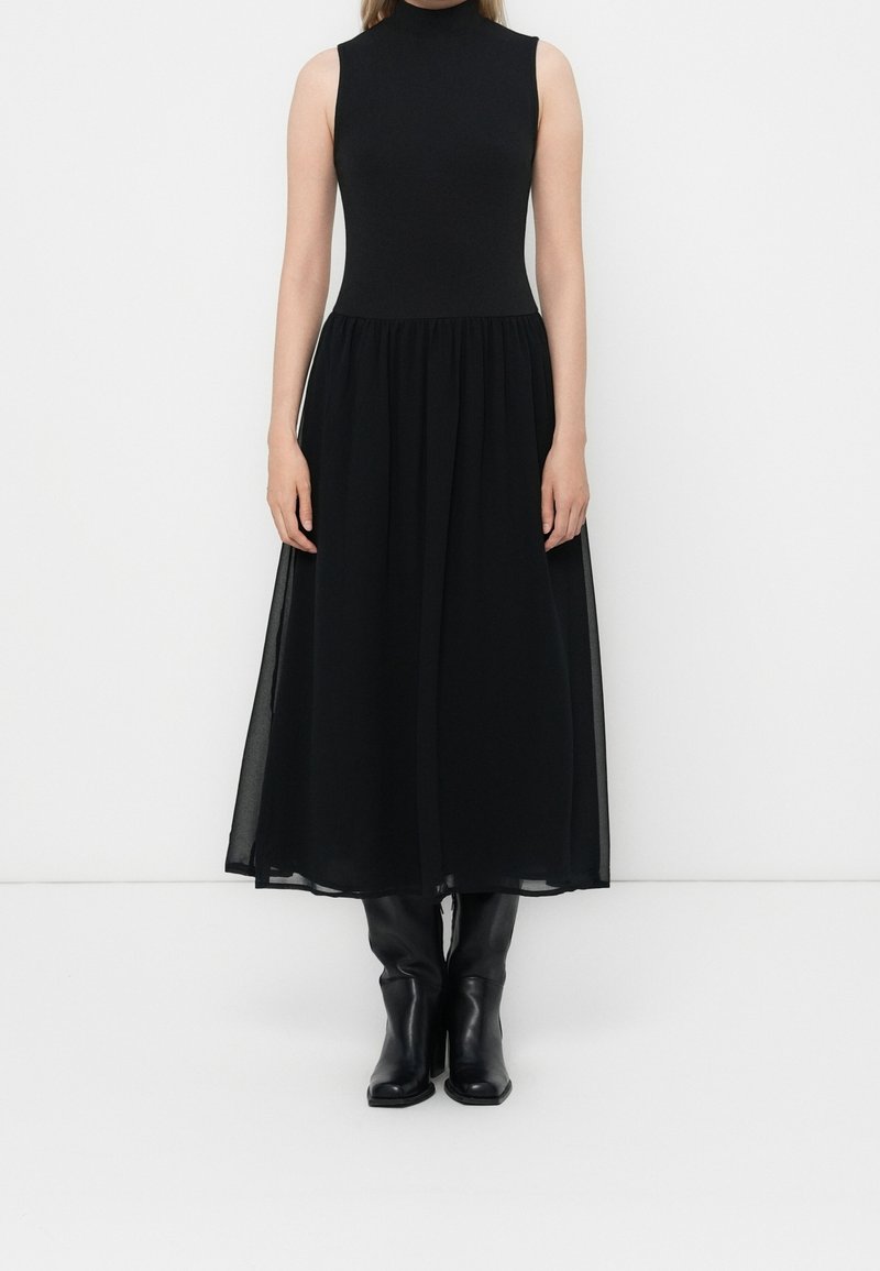 Femme portant une robe noire sans manches à col roulé avec une jupe midi transparente et des bottes noires montantes, debout sur fond blanc.