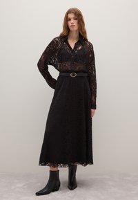 Donna che indossa una camicia nera in pizzo a maniche lunghe, una gonna midi nera in pizzo coordinata, una cintura nera e stivaletti neri alla caviglia, in piedi contro un muro semplice.