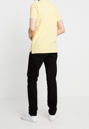 Mężczyzna stoi tyłem do kamery, ubrany w żółtą koszulkę z krótkim rękawem, czarne dżinsy i białe trampki, na jednolitym białym tle.