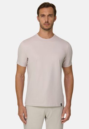 Man met kort bruin haar en lichte baard, die een effen lichtbeige T-shirt met korte mouwen en bijpassende lichtbeige broek draagt, kijkt naar voren.