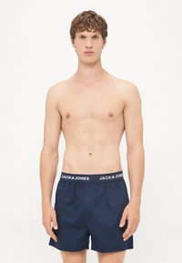 Marineblå svømmedrakter med elastisk talje og hvid "JACK & JONES" branding. Glat tekstur, afslappet pasform, over knæ længde.
