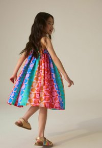 Jeune fille tournant dans une robe sans manches colorée avec des motifs de coquillages et portant des sandales dorées sur un fond uni.
