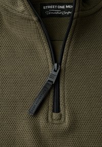 Olivově zelený svrchní zipový top vyrobený z texturovaného waffle pleteného materiálu. Obsahuje černý zip s táhněte značkou "SEEK ADVENTURE" a značkovou nášivkou.