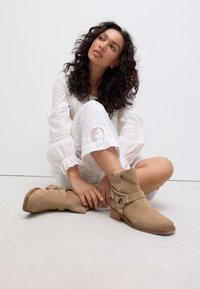 Beige Wildleder-Stiefeletten mit kurzem Blockabsatz, ausgestattet mit Riemen- und Schnallendetails, vor einem neutralen Hintergrund mit einem lässigen Outfit.