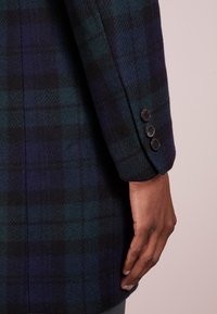 Cappotto in lana a quadri verde scuro e blu navy, con una vestibilità sartoriale, dettagli con polsini a tre bottoni e una morbida texture spazzolata.