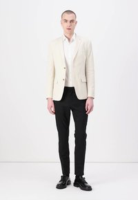 Lichtbeige blazer met twee voorknopen, gecombineerd met een wit overhemd en zwarte broek. Zwarte leren schoenen maken de outfit compleet.