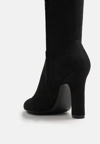 Botte à talon haut en cuir suedé noir avec une finition lisse, un bout arrondi et une fermeture éclair sur le côté. Le talon a une conception solide et légèrement courbée.