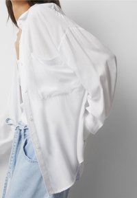 Camicia bianca a maniche lunghe realizzata in tessuto leggero, con tasca, bottoni e vestibilità comoda, abbinata a jeans denim azzurri.