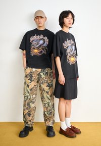 Black graphic t-shirts with a colorful scorpion design, paired with camouflage pants and a black pleated skirt. Black and brown shoes worn.