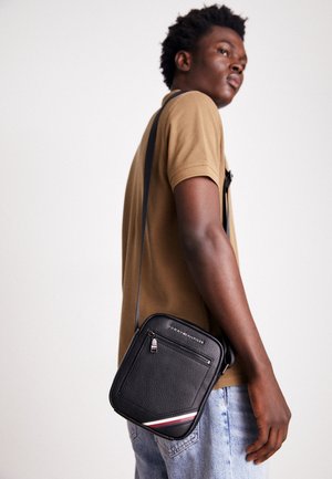 Jeune homme portant une chemise marron et un jean clair, porte un sac bandoulière noir Tommy Hilfiger avec une poche zippée à l'avant et un logo.