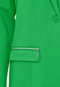 Chaqueta verde con un bolsillo con cremallera en el frente. La tela es texturizada y la cremallera es plateada con una pequeña lengüeta.