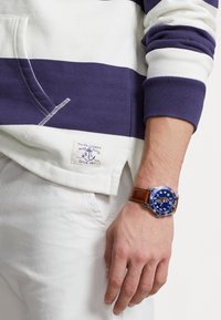 Reloj azul con esfera redonda, marcas blancas y correa de cuero marrón, combinado con un sudadera a rayas azul marino y blanca.