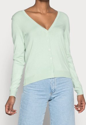 Donna che indossa un cardigan verde chiaro con scollo a V e bottoni, maniche lunghe e jeans a vita alta azzurri.