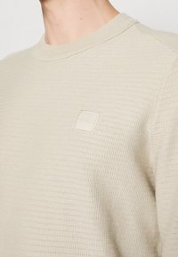 Pull en tricot beige texturé avec col rond, doté d'un petit patch logo carré portant l'inscription "BOSS" sur le côté gauche de la poitrine.