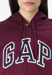 Close-up of a person wearing a burgundy GAP hoodie with a front zipper and white-bordered navy letters on the chest.