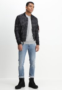 Giacca bomber in camouflage nera sopra una felpa grigia, abbinata a jeans azzurri chiari e stivali neri con lacci. Presenta polsini e orlo a costine.