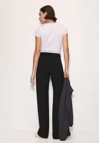 Čierne nohavice s vysokým pásom v kombinácii s priliehavým sivým tričkom. Modelka drží tmavú bundu v pravej ruke a stojí na jednoduchom pozadí.