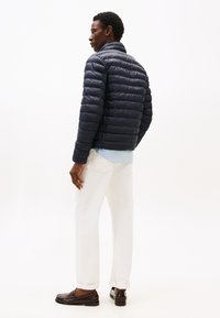 Doudoune matelassée bleu marine avec col montant, portée sur une chemise à rayures bleu clair, associée à un pantalon blanc et des mocassins marron.