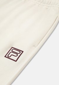 Beige jogginghousut, jotka on valmistettu pehmeästä kankaasta ja joissa on joustava vyötärö. Vasemmalla puolella lähellä taskua on burgundinvärinen kirjottu logo.