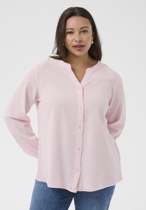 Vrouw met lang donker haar die een lichtroze blouse met knopen en een blauwe spijkerbroek draagt, staand tegen een effen lichte achtergrond.